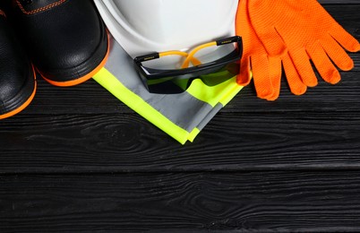 Photo of Pair of working boots, hard hat, protective gloves, reflective vest and goggles on black wooden background, flat lay. Space for text