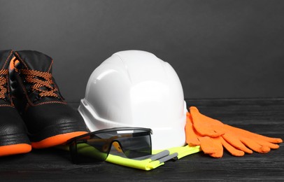 Photo of Pair of working boots, hard hat, protective gloves, reflective vest and goggles on black wooden surface against gray background