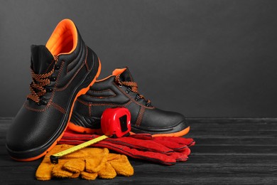 Photo of Pair of working boots, protective gloves and tape measure on black wooden surface against gray background, space for text