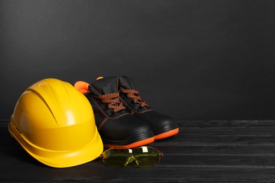 Photo of Pair of working boots, hard hat and goggles on black wooden surface, space for text