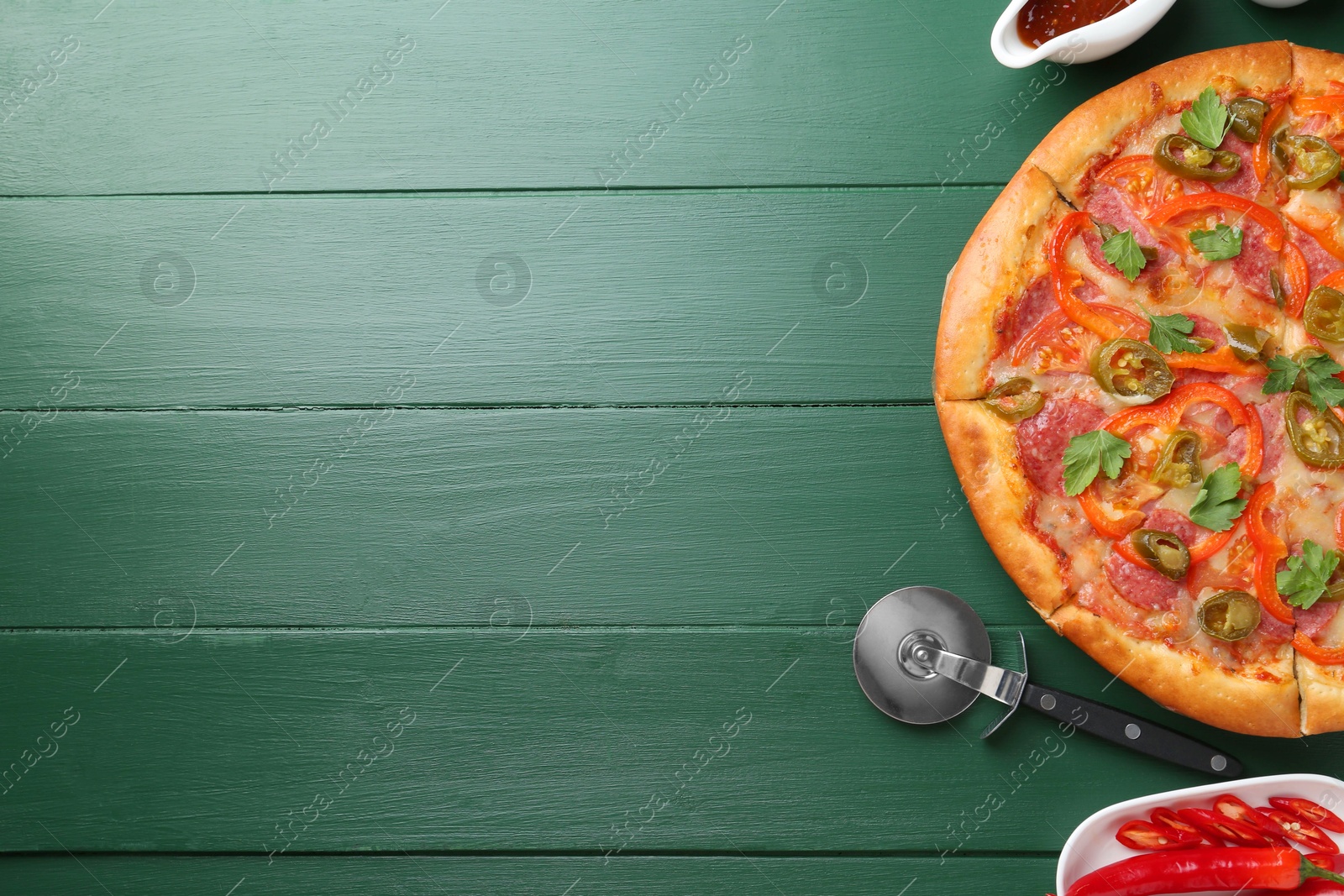 Photo of Delicious pizza Diablo, sauce, fresh chili peppers and cutter on green wooden table, flat lay. Space for text