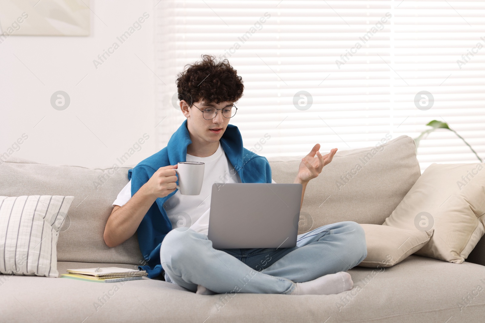 Photo of Teenager holding cup of drink working with laptop on sofa at home. Remote job