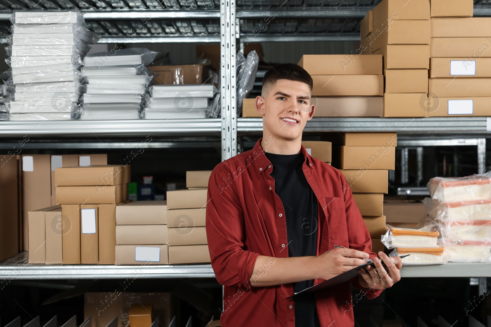 Photo of Smiling young man with clipboard near cardboard boxes in auto store