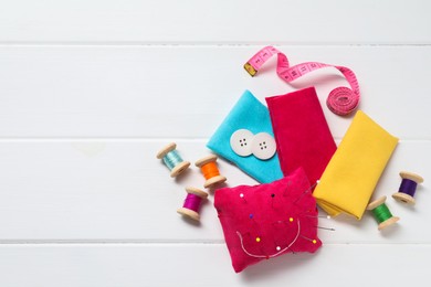 Red pincushion with pins and other sewing tools on white wooden table, flat lay. Space for text