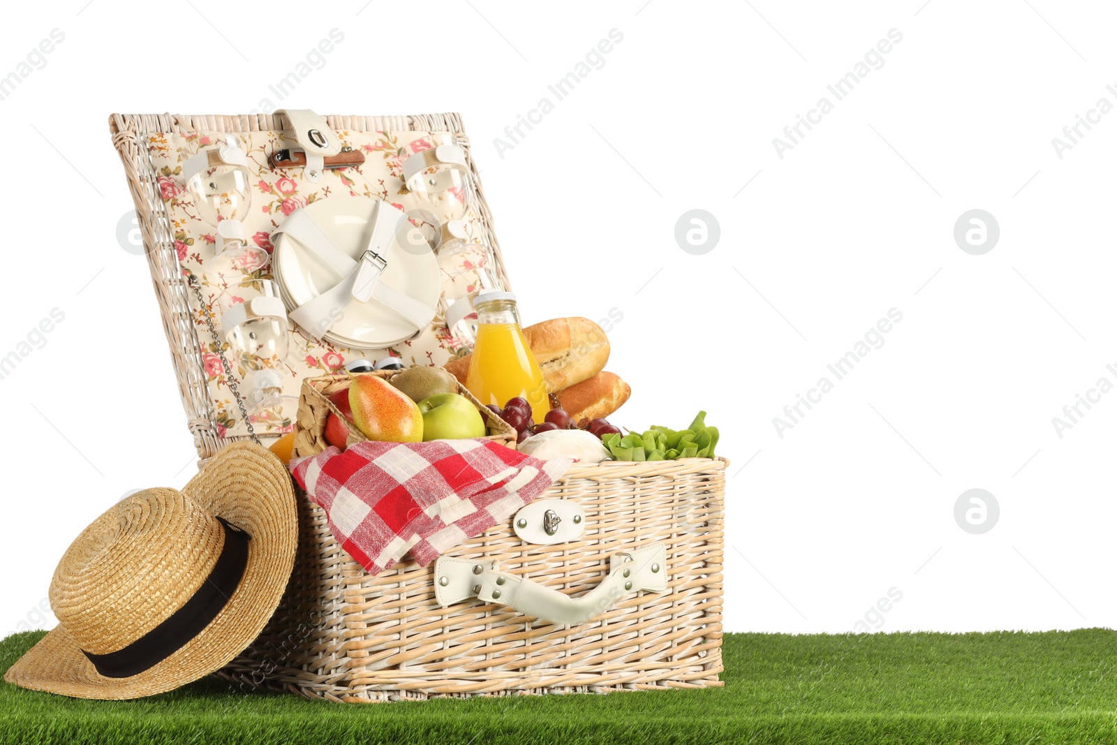 Photo of Picnic wicker basket with food, tableware, bottle of juice and hat on grass against white background. Space for text