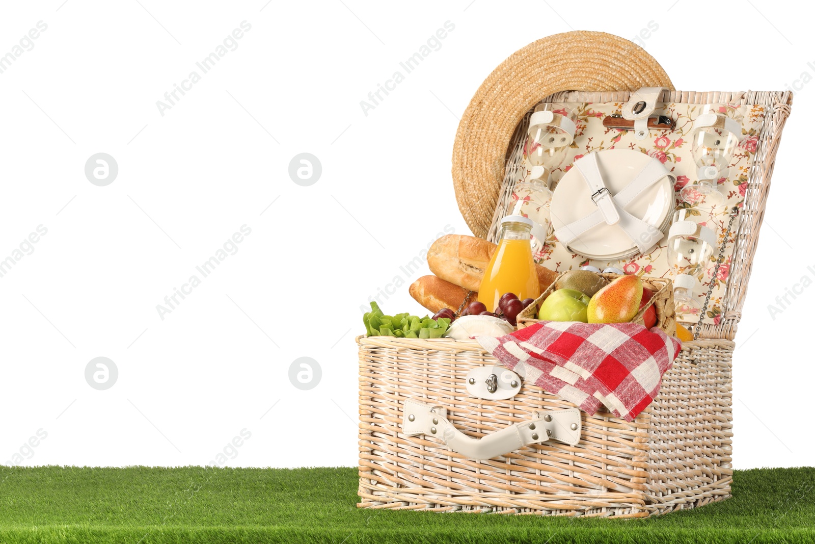 Photo of Picnic wicker basket with food, tableware, bottle of juice and hat on grass against white background. Space for text