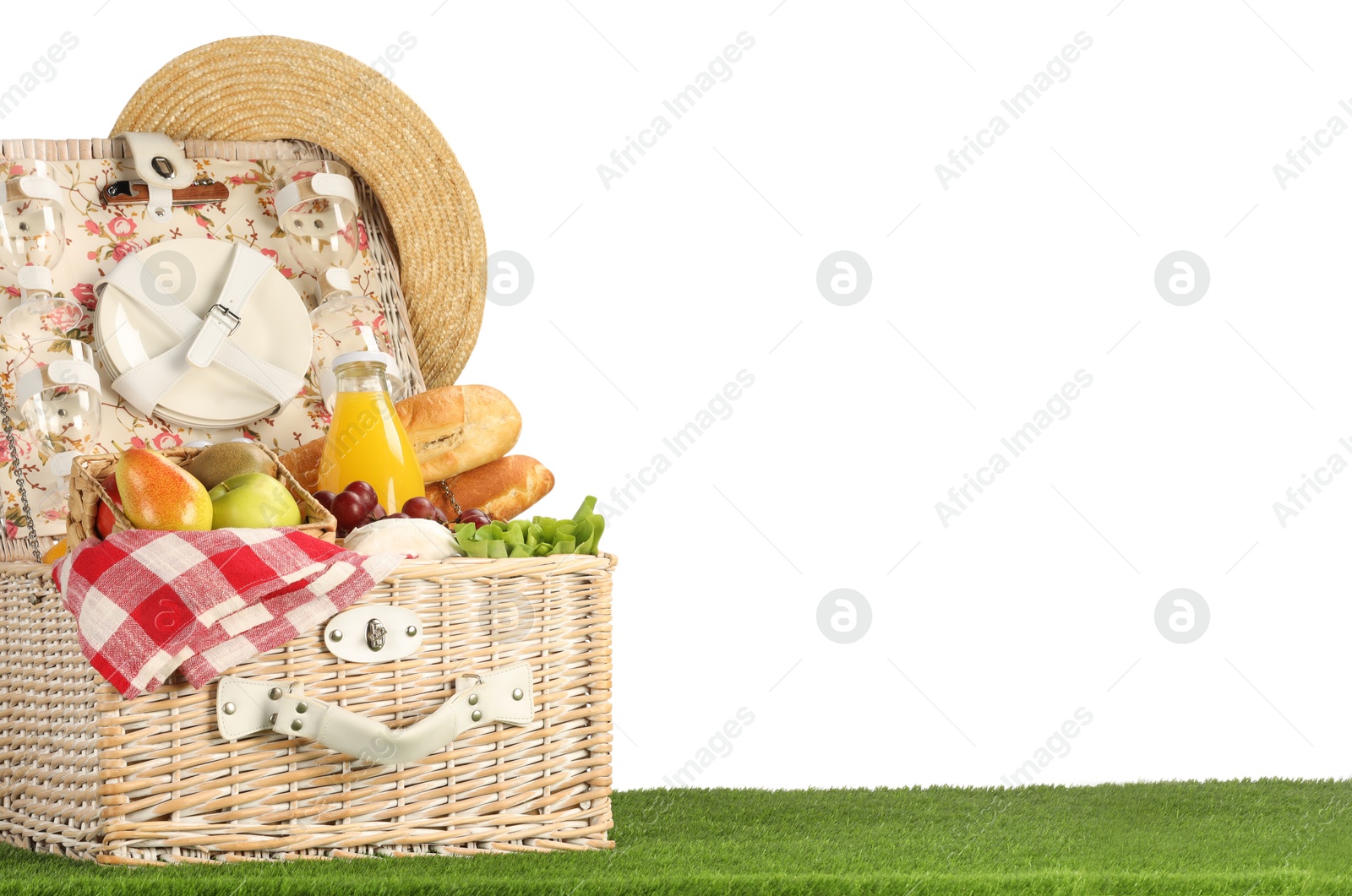 Photo of Picnic wicker basket with food, tableware, bottle of juice and hat on grass against white background. Space for text