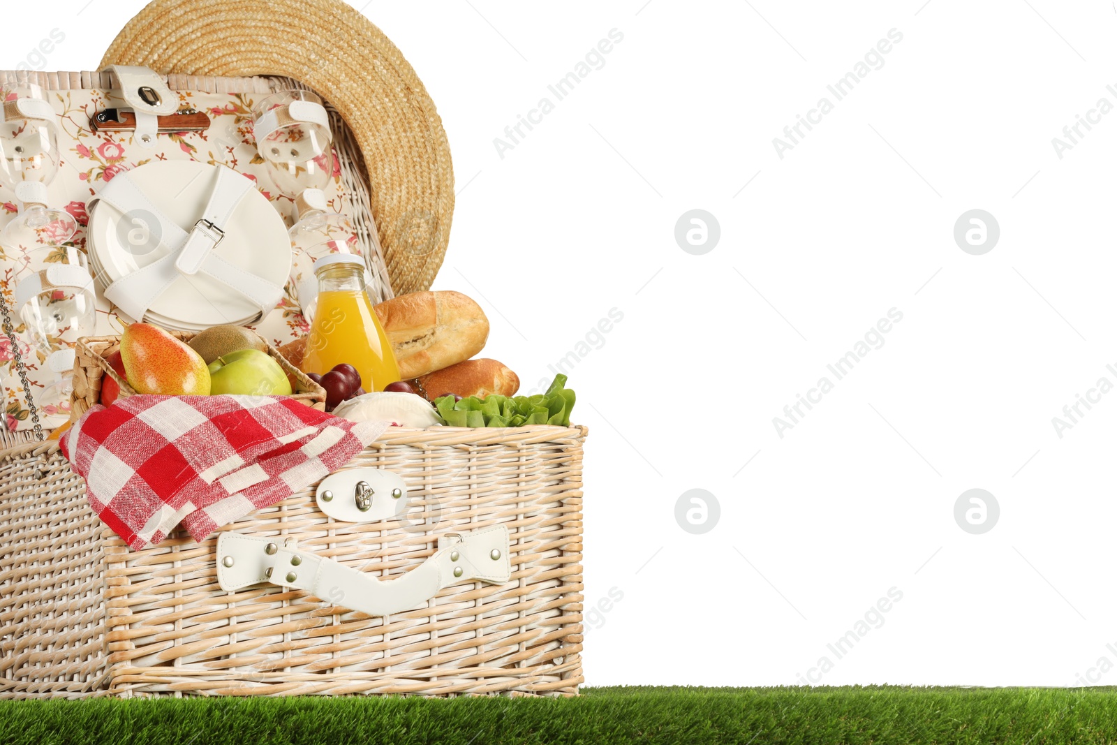 Photo of Picnic wicker basket with food, tableware, bottle of juice and hat on grass against white background. Space for text