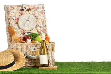 Photo of Picnic wicker basket with food, tableware, bottle of wine and hat on grass against white background. Space for text