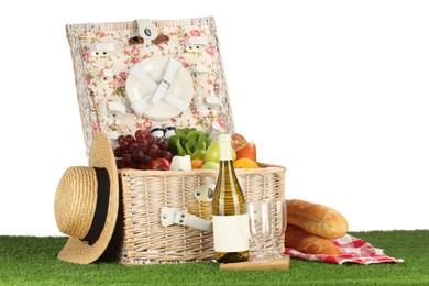 Picnic wicker basket with food, tableware, bottle of wine and hat on grass against white background