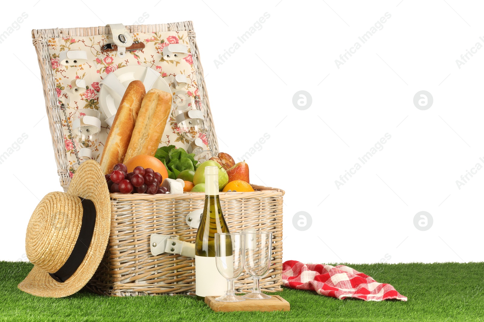 Photo of Picnic wicker basket with food, tableware, bottle of wine and hat on grass against white background. Space for text