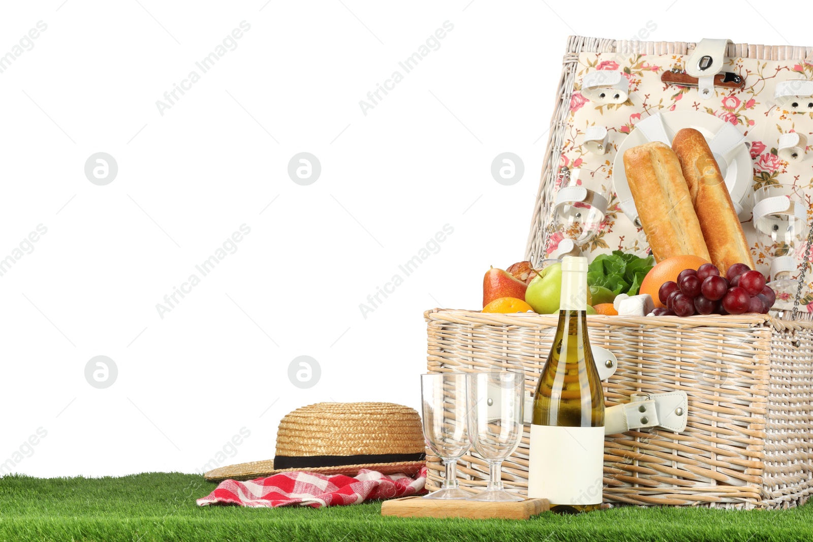 Photo of Picnic wicker basket with food, tableware, bottle of wine and hat on grass against white background. Space for text