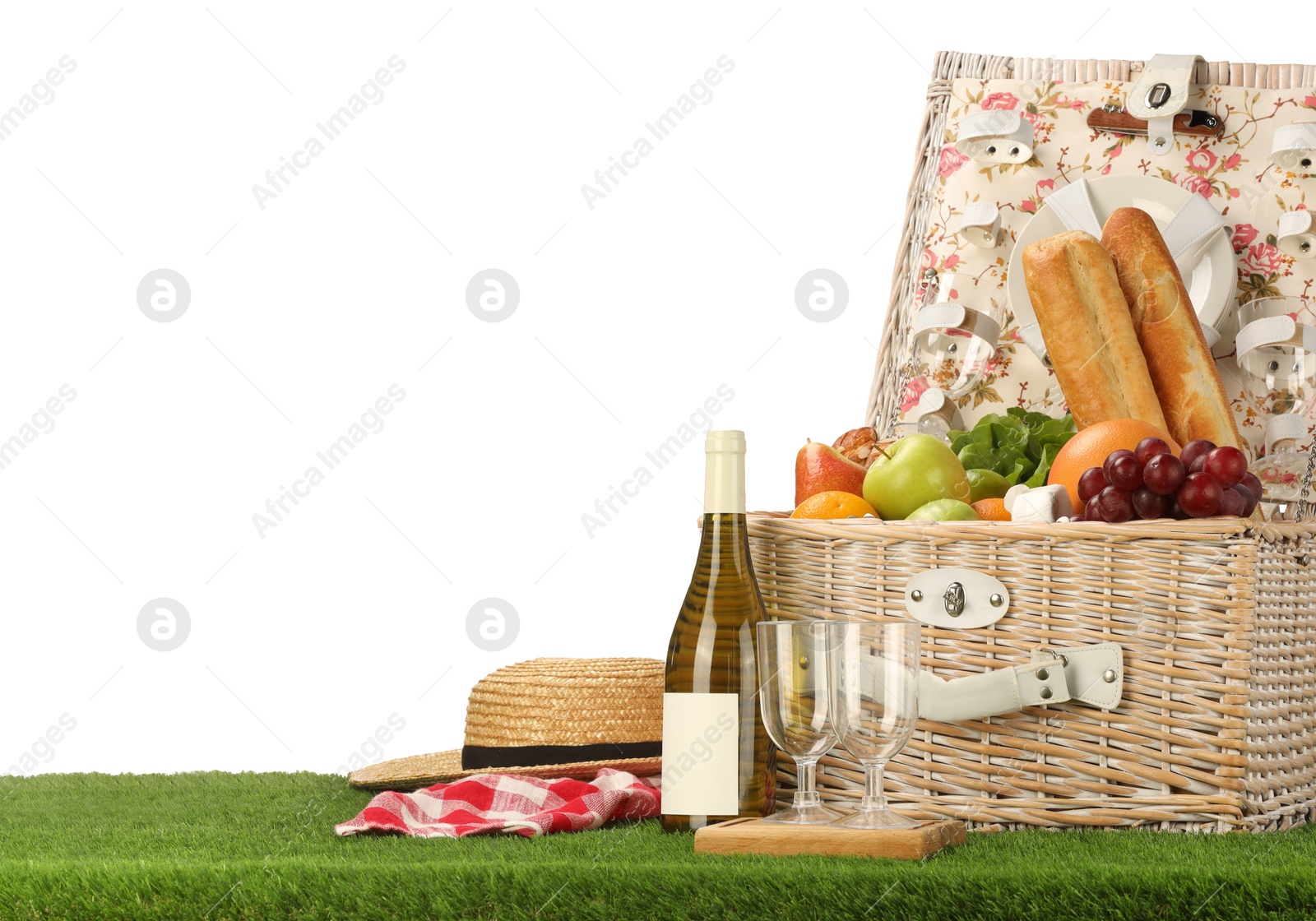 Photo of Picnic wicker basket with food, tableware, bottle of wine and hat on grass against white background. Space for text