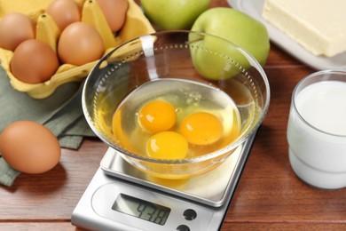 Kitchen scale with bowl of raw eggs and milk on wooden table, closeup