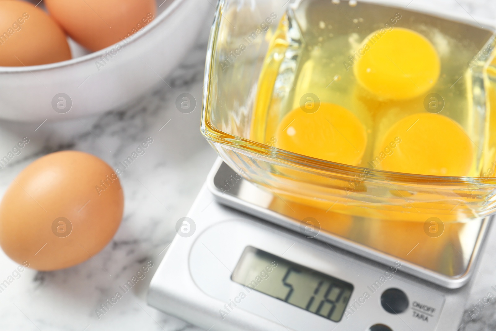 Photo of Kitchen scale with bowl of raw eggs on white marble table, closeup