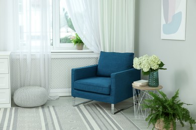 Soft armchair, side table, flowers and window with curtains in room. Interior design