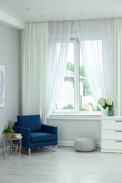 Soft armchair, side table, flowers and window with curtains in room. Interior design