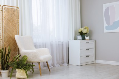 Soft armchair, chest of drawers, houseplants and window with curtains in room. Interior design