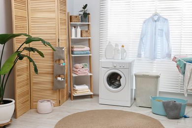 Photo of Washing machine, detergents, towels, baskets and folding screen in laundry room