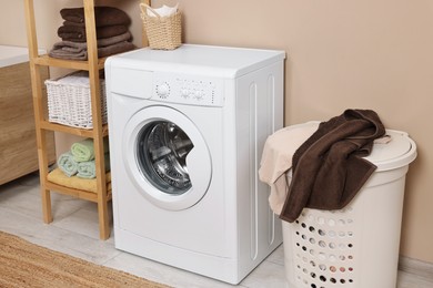 Photo of Washing machine, basket and shelving unit near beige wall indoors