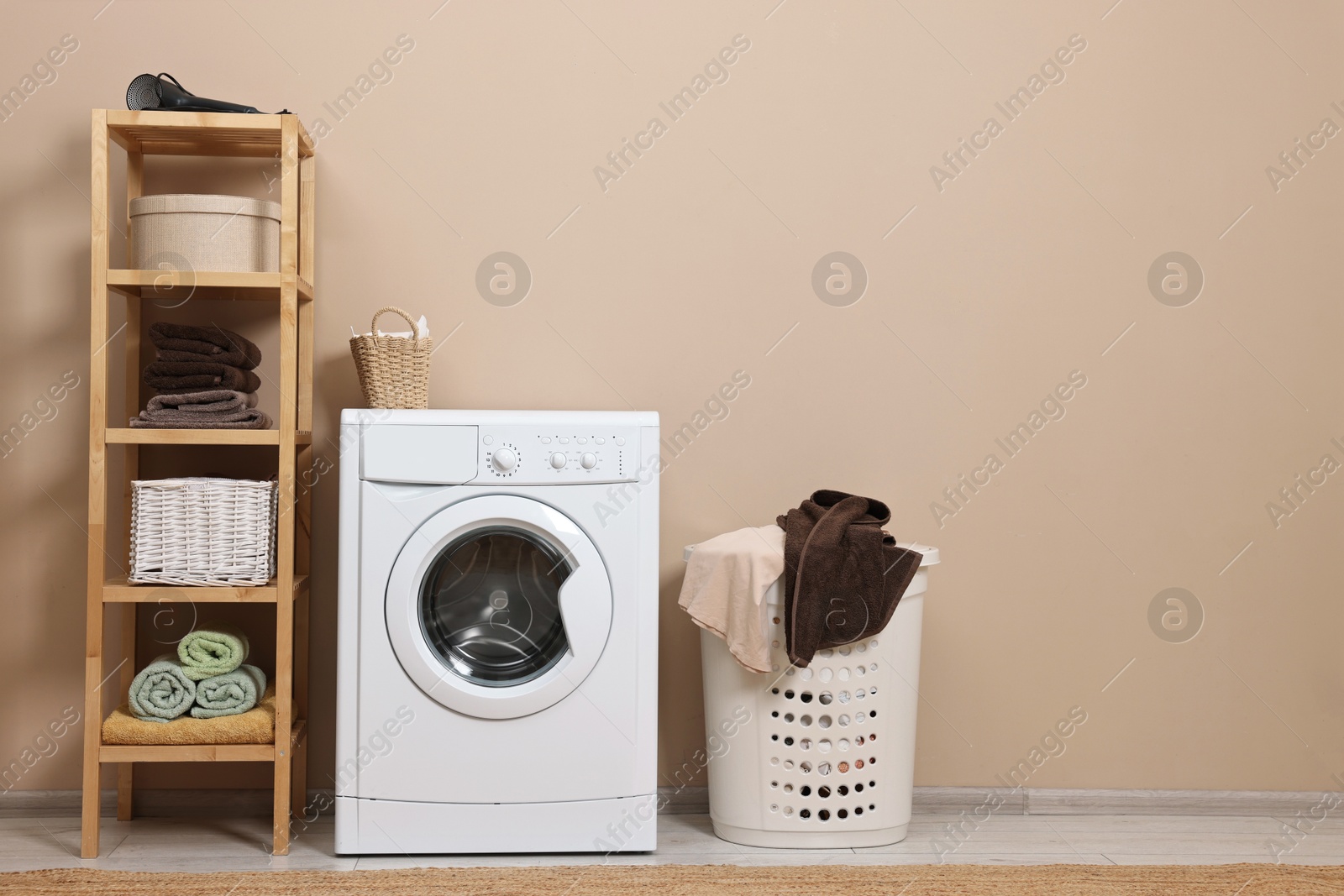 Photo of Washing machine, basket and shelving unit near beige wall indoors. Space for text