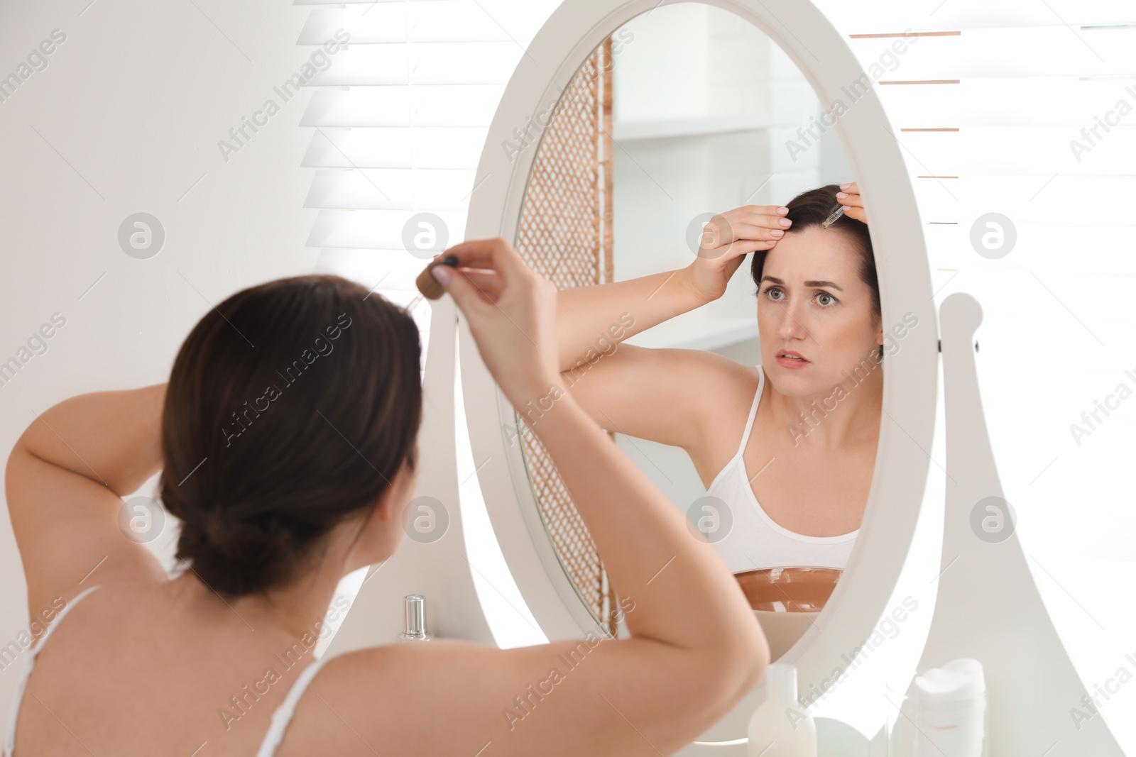 Photo of Hair loss problem. Woman applying serum onto hairline near mirror at home