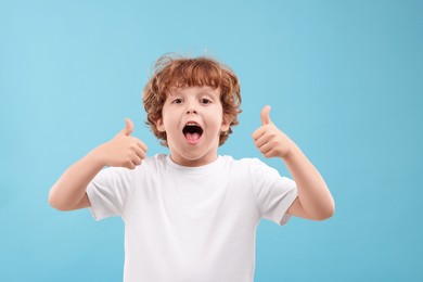Photo of Portrait of emotional little boy showing thumbs up on light blue background