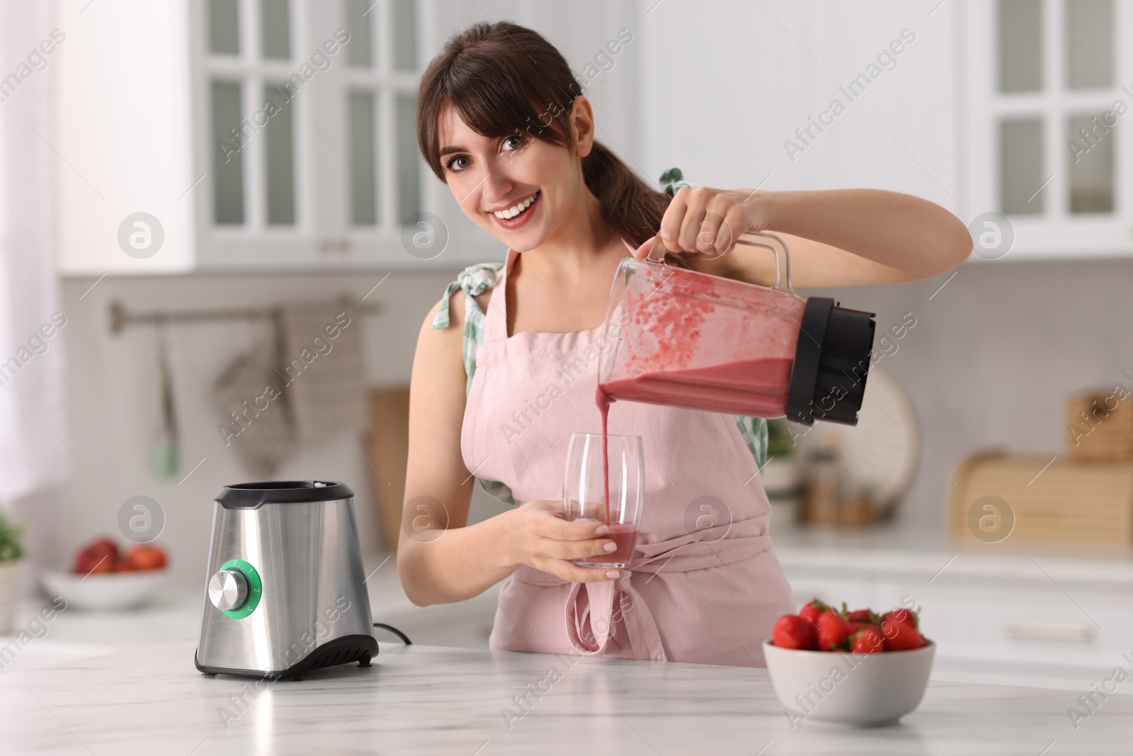 Photo of Young woman pouring fresh smoothie from blender cup into glass at white marble table in kitchen