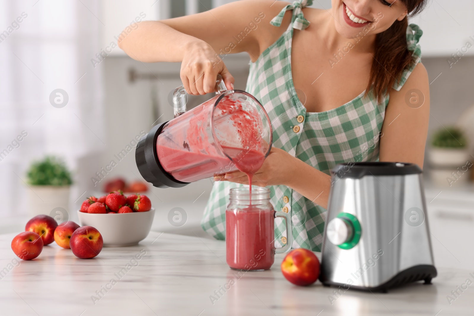 Photo of Woman pouring fresh smoothie from blender cup into glass at white marble table in kitchen, closeup