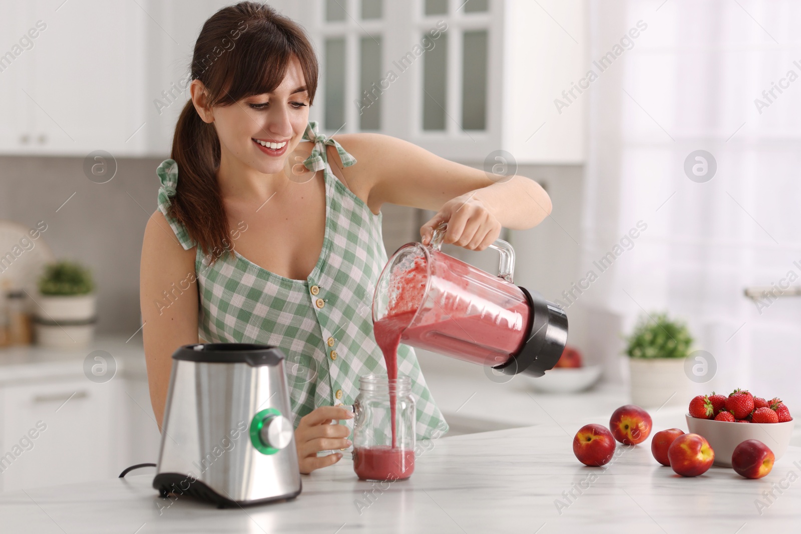 Photo of Young woman pouring fresh smoothie from blender cup into glass at white marble table in kitchen