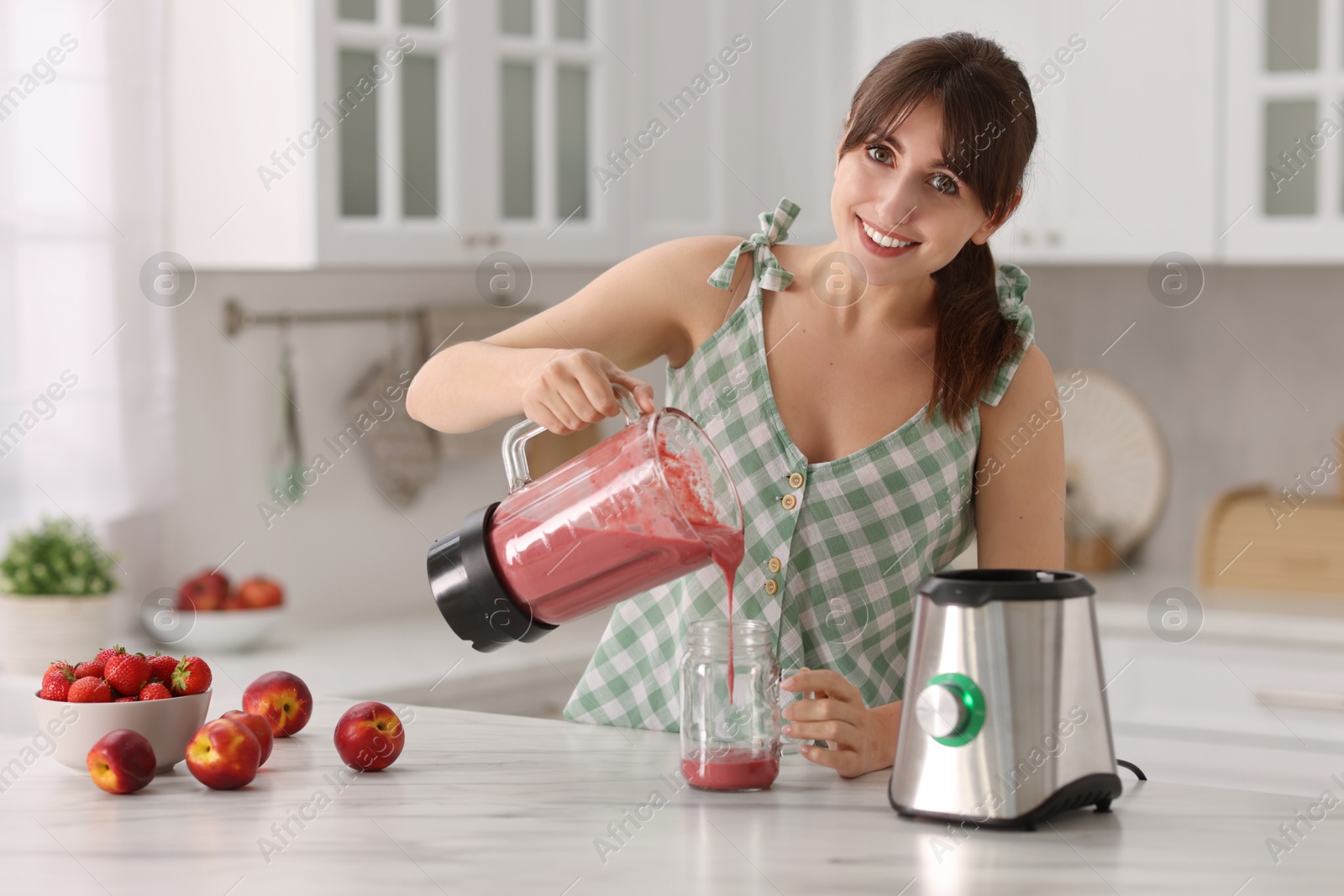 Photo of Young woman pouring fresh smoothie from blender cup into glass at white marble table in kitchen