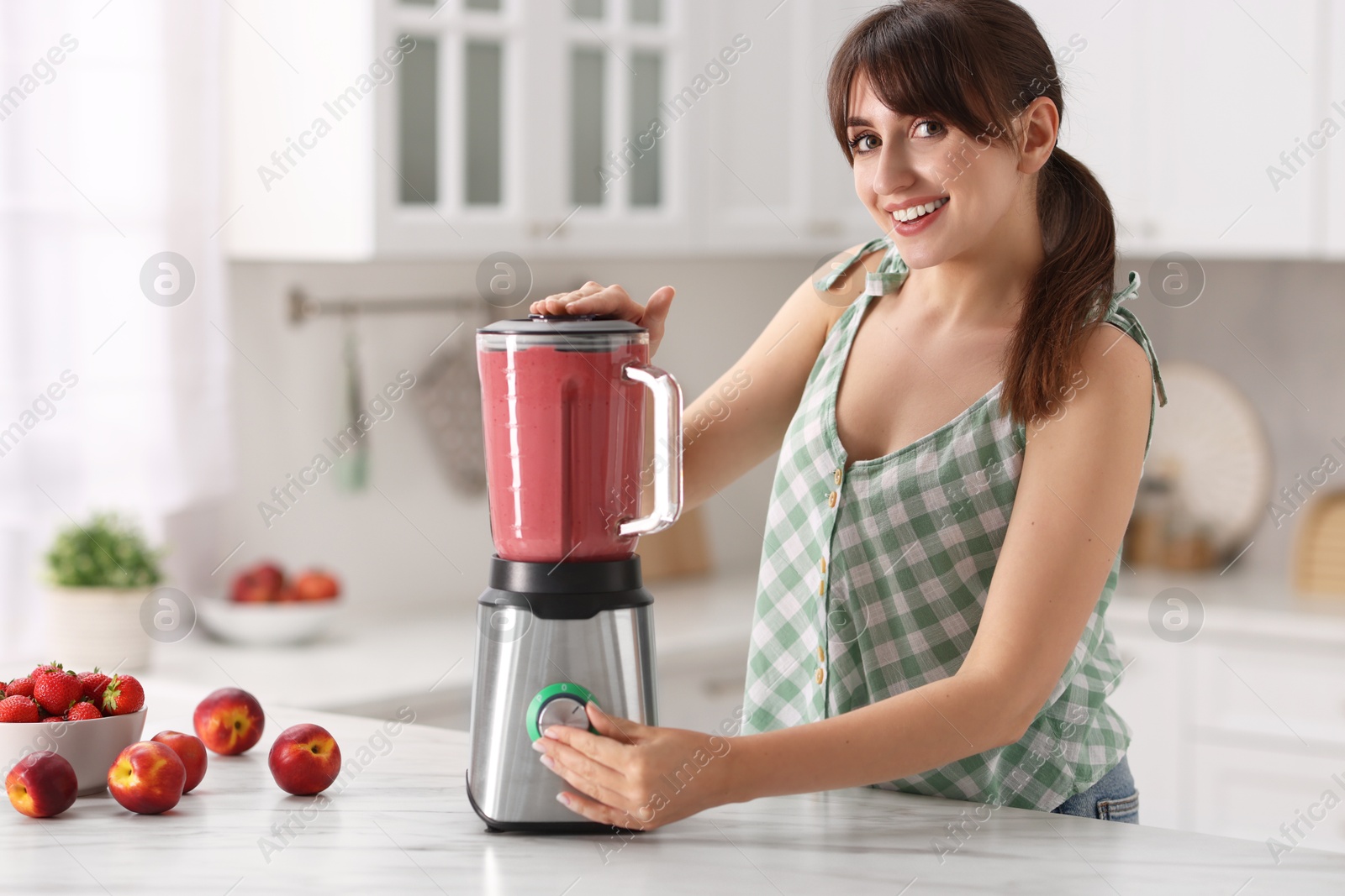 Photo of Young woman making delicious smoothie with blender at white marble table in kitchen