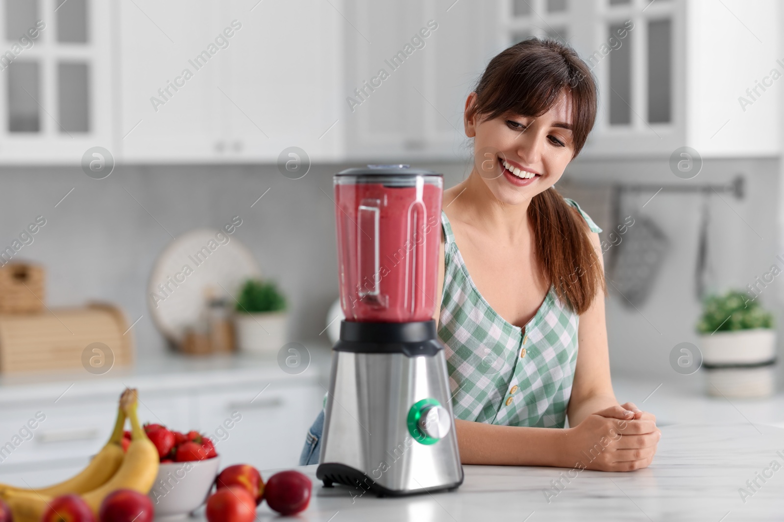 Photo of Young woman making delicious smoothie with blender at white marble table in kitchen