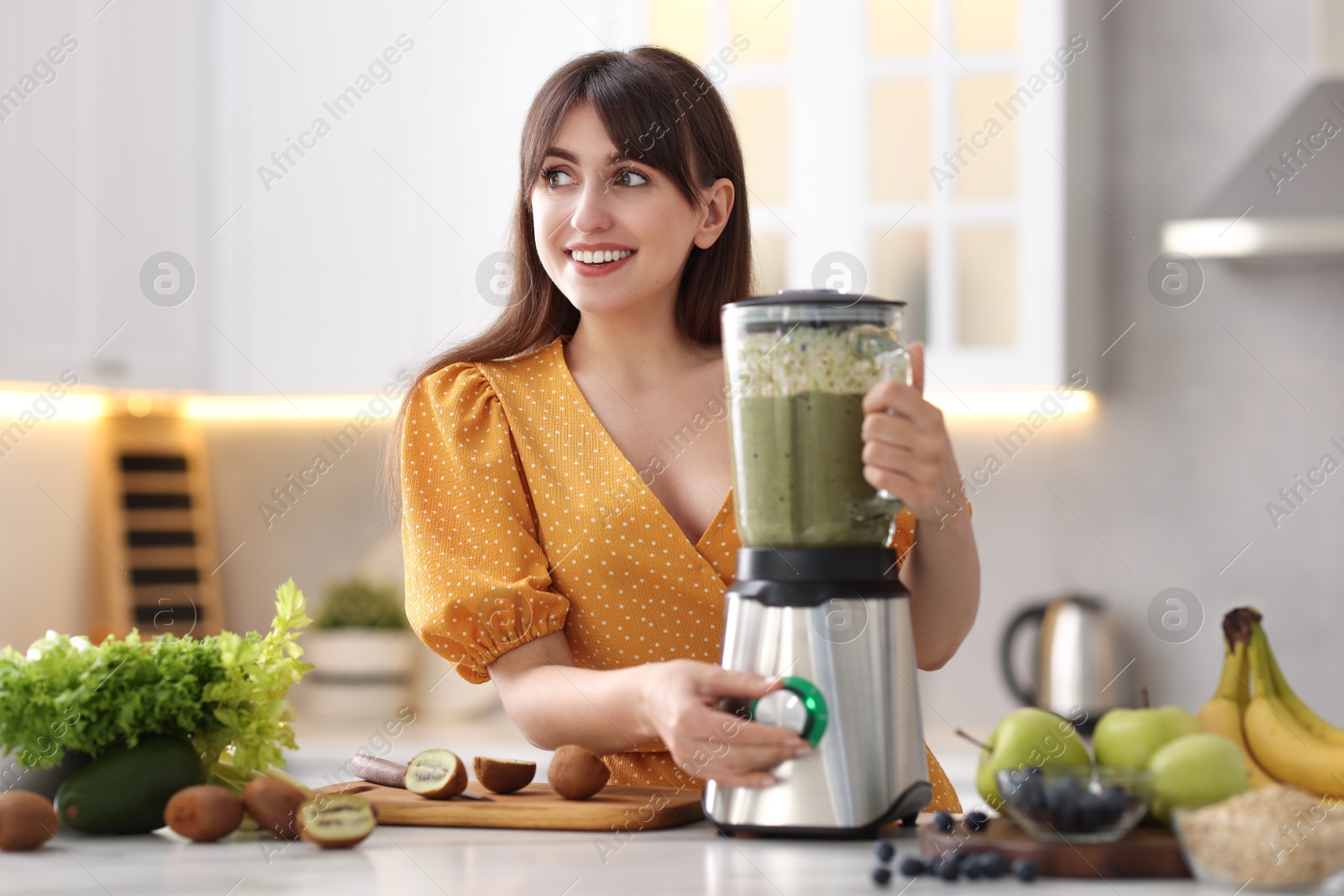 Photo of Young woman making delicious smoothie with blender at white marble table in kitchen