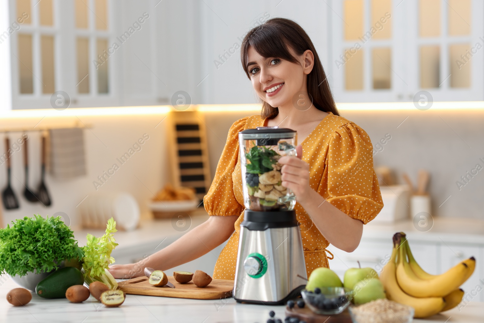 Photo of Young woman making delicious smoothie with blender at white marble table in kitchen