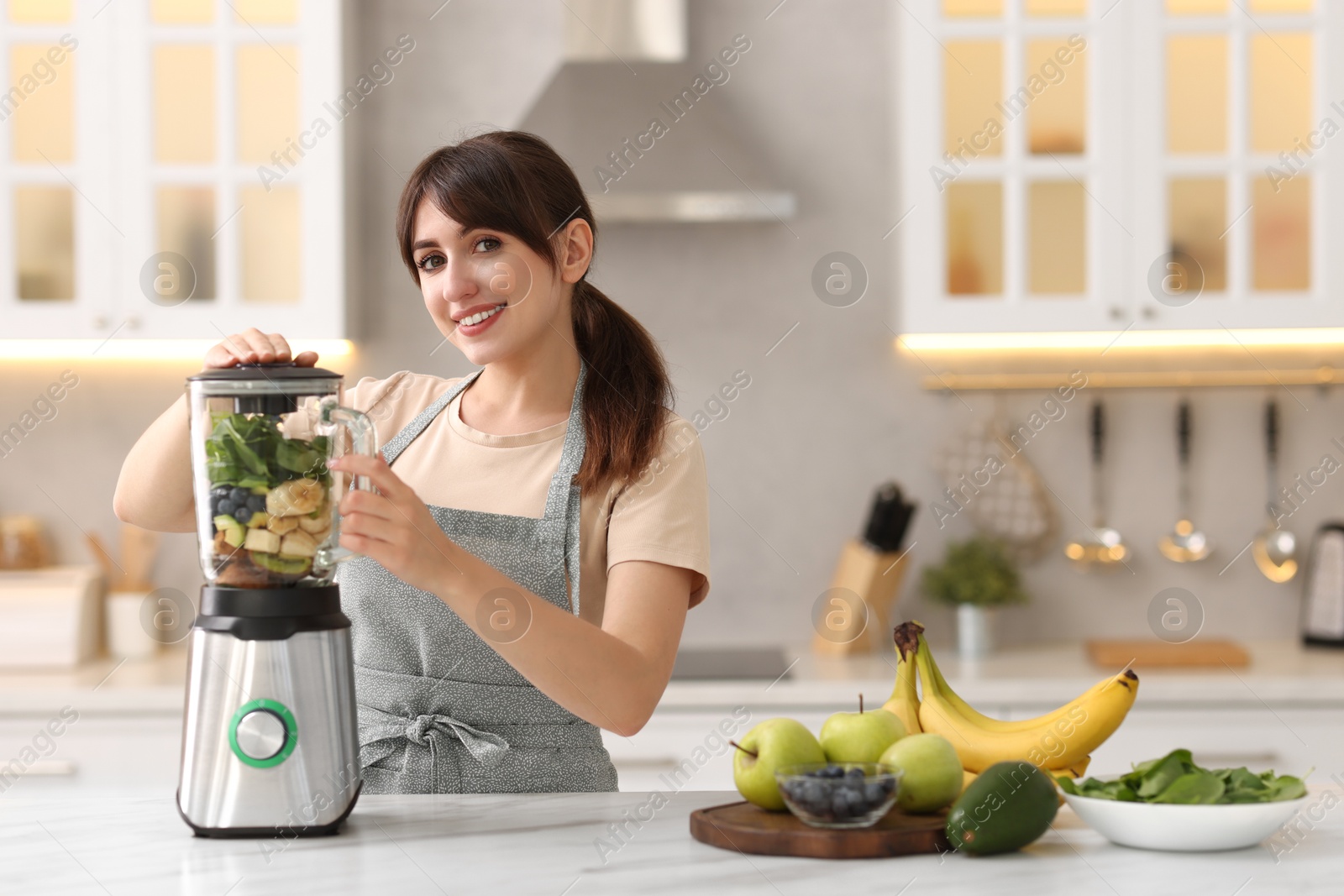 Photo of Young woman making delicious smoothie with blender at white marble table in kitchen