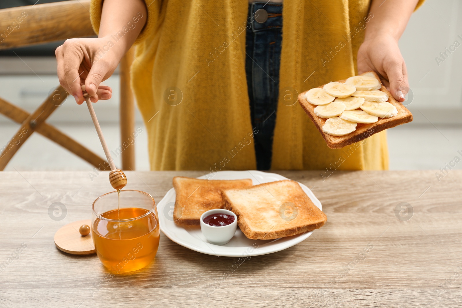 Photo of Woman pouring honey onto toast with banana at table indoors, closeup