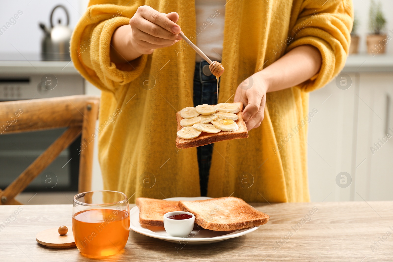 Photo of Woman pouring honey onto toast with banana at table indoors, closeup