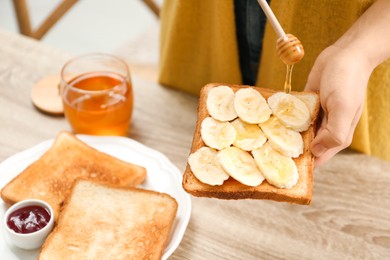 Photo of Woman pouring honey onto toast with banana at table indoors, closeup