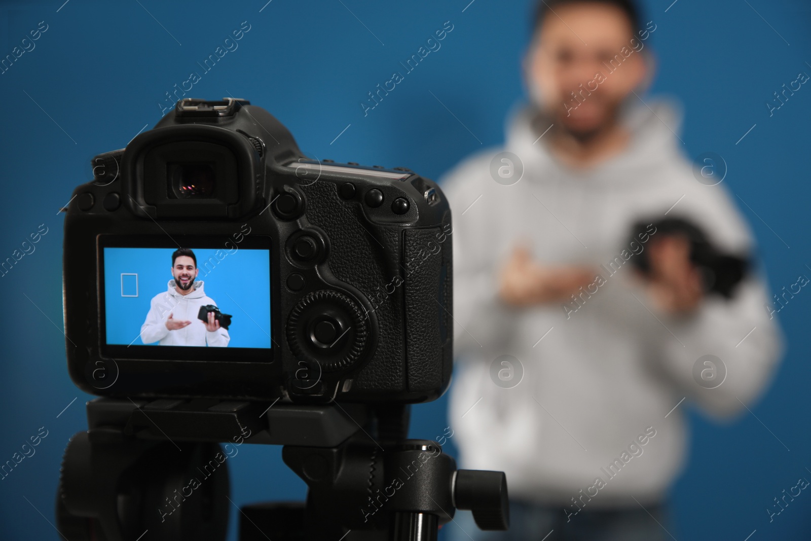 Photo of Young blogger with camera shooting video against blue background, focus on screen