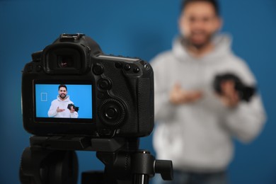 Young blogger with camera shooting video against blue background, focus on screen