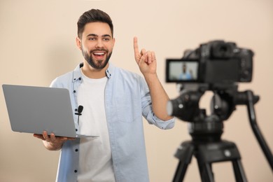 Young blogger with laptop recording video on camera against beige background