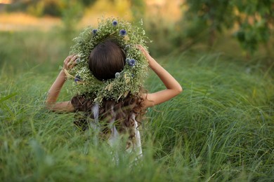 Photo of Little girl wearing wreath made of beautiful flowers in field, back view