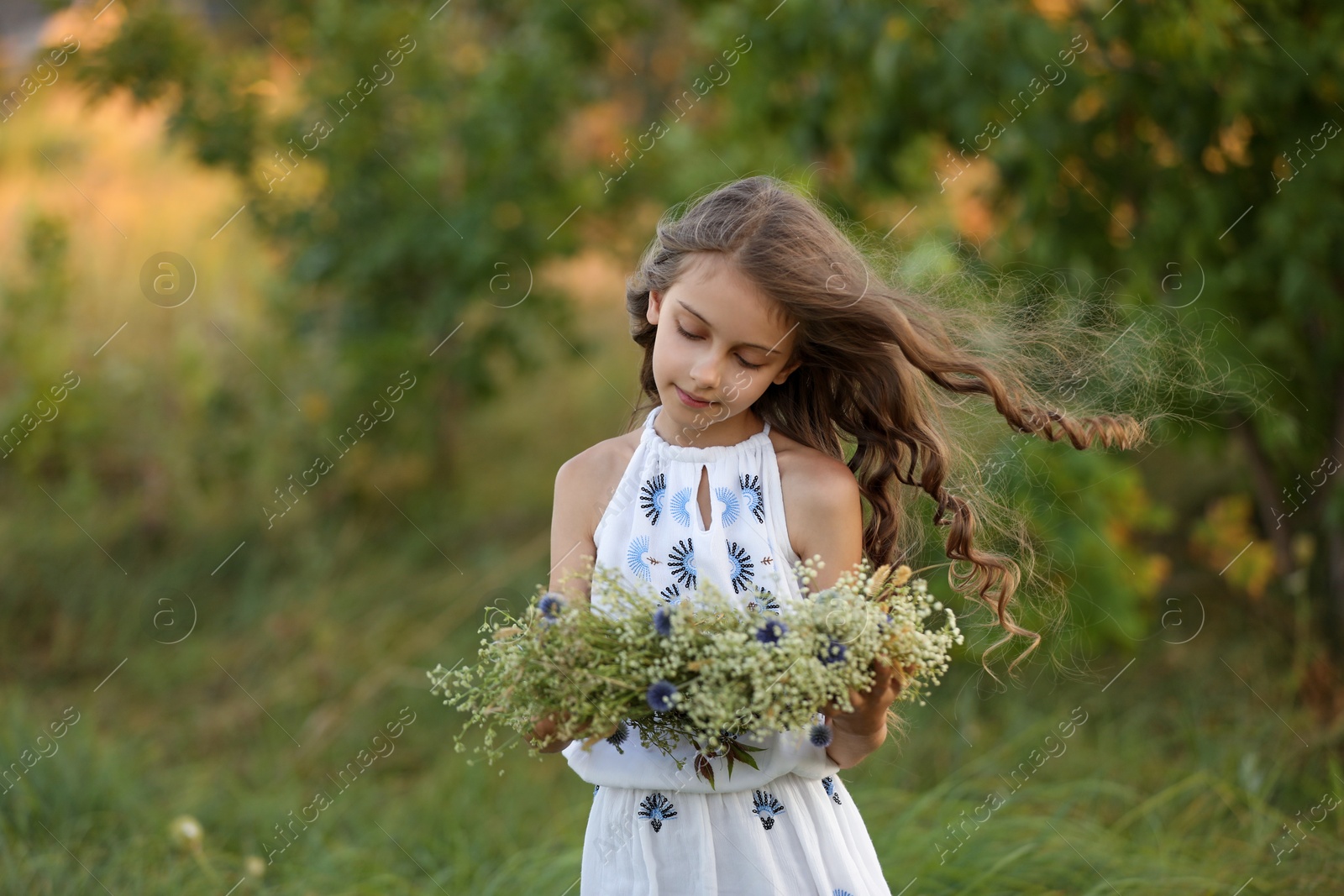 Photo of Cute little girl holding wreath made of beautiful flowers in field