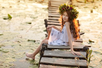 Photo of Cute little girl wearing wreath made of beautiful flowers on pier near pond