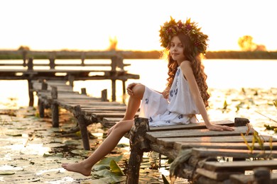 Photo of Cute little girl wearing wreath made of beautiful flowers on pier near river at sunset