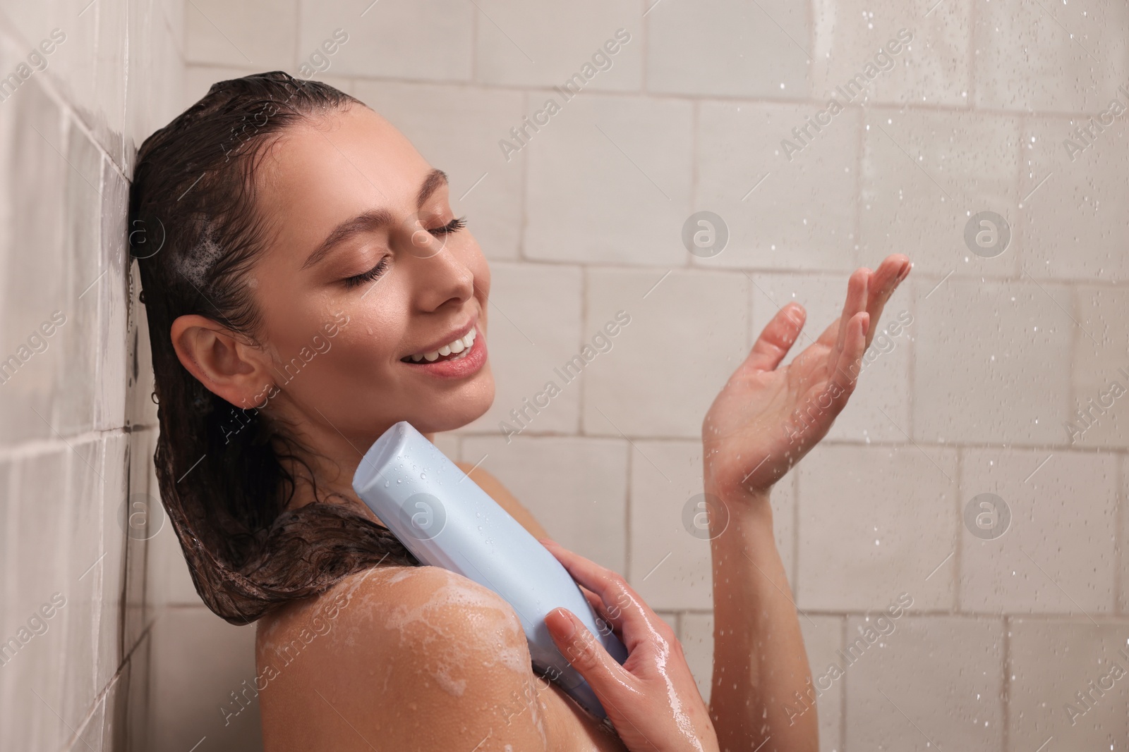 Photo of Happy woman with bottle of shampoo in shower at home. Washing hair