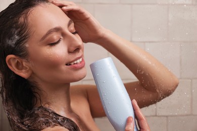 Photo of Happy woman with bottle of shampoo in shower at home. Washing hair