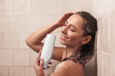 Photo of Happy woman with bottle of shampoo in shower at home. Washing hair