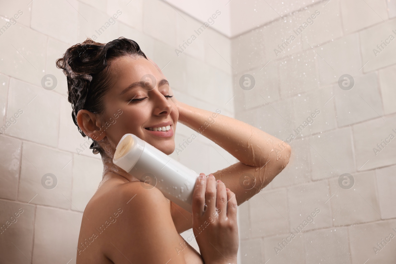 Photo of Happy woman with bottle of shampoo in shower at home. Washing hair